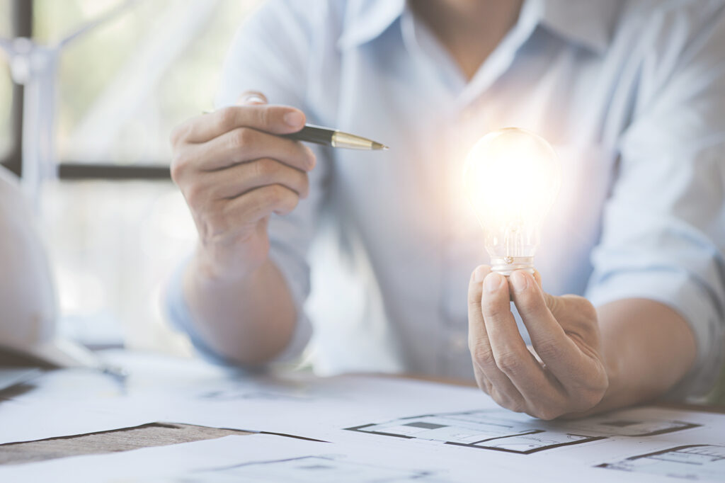 Person mit Kugelschreiber und Glühbirne in der Hand, vor einem Schreibtisch mit Grundrissen eines Bauprojektes